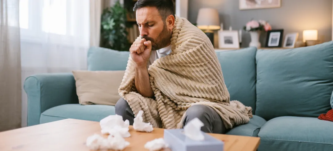 Hombre enfermo, abrigado con una manta y tosiendo en el salón de su casa